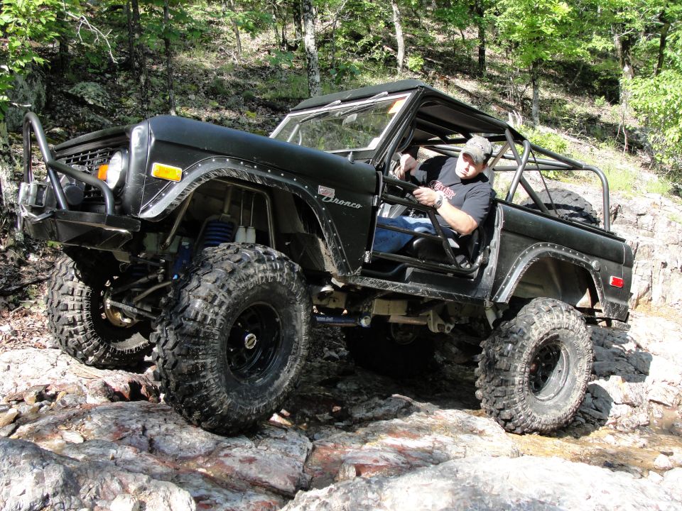 Black Ford Bronco with large tires driving over rocks on a wooded trail