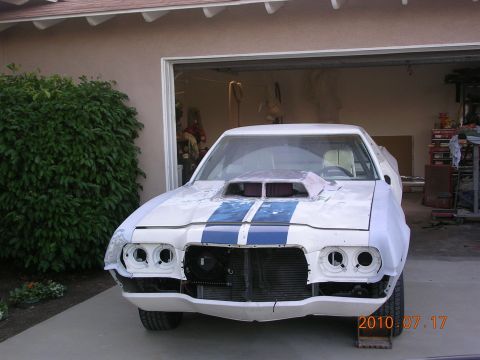 1972 Ford Ranchero - A white 1972 Ford Ranchero with racing stripes and a hood scoop is shown in a three-quarter front view, appearing to be under restoration.