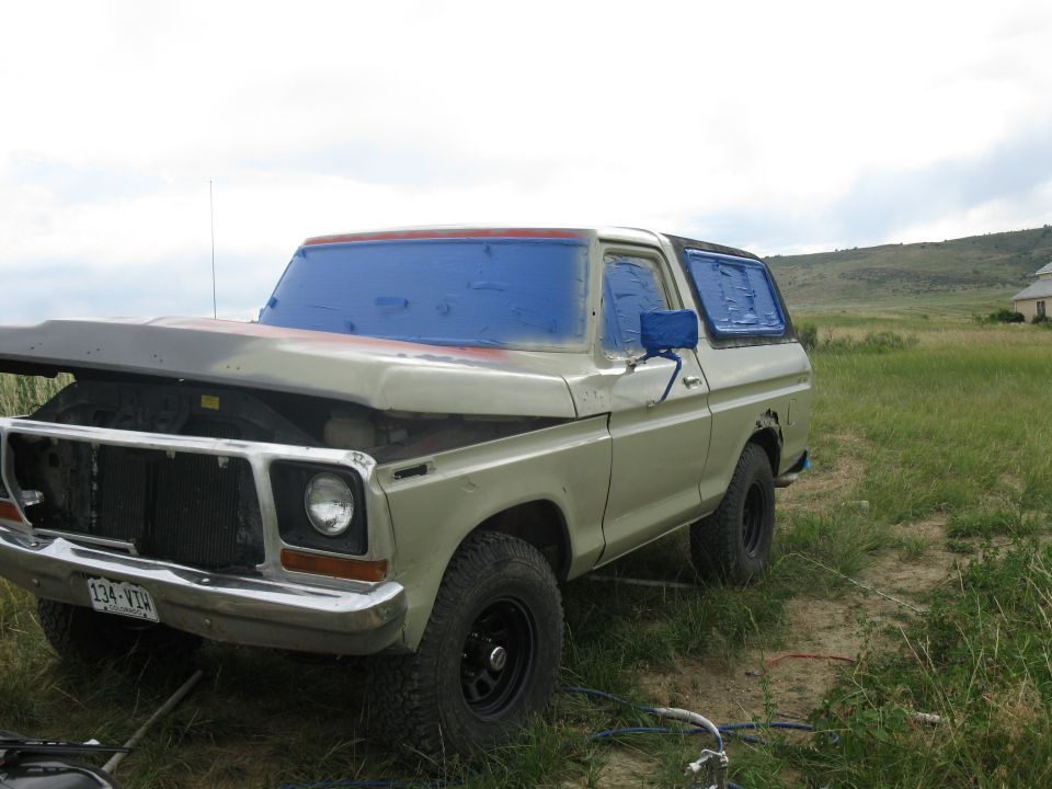 Tan 1978 Ford Bronco with hood open and windows taped, parked in a grassy field