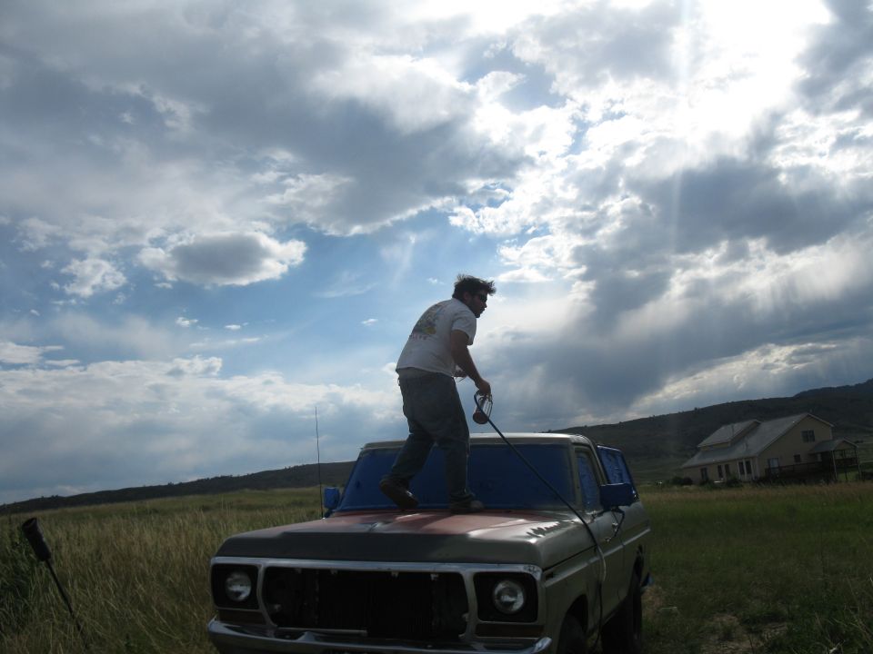 Man standing on the hood of a vintage truck, holding a rope, with a cloudy sky overhead