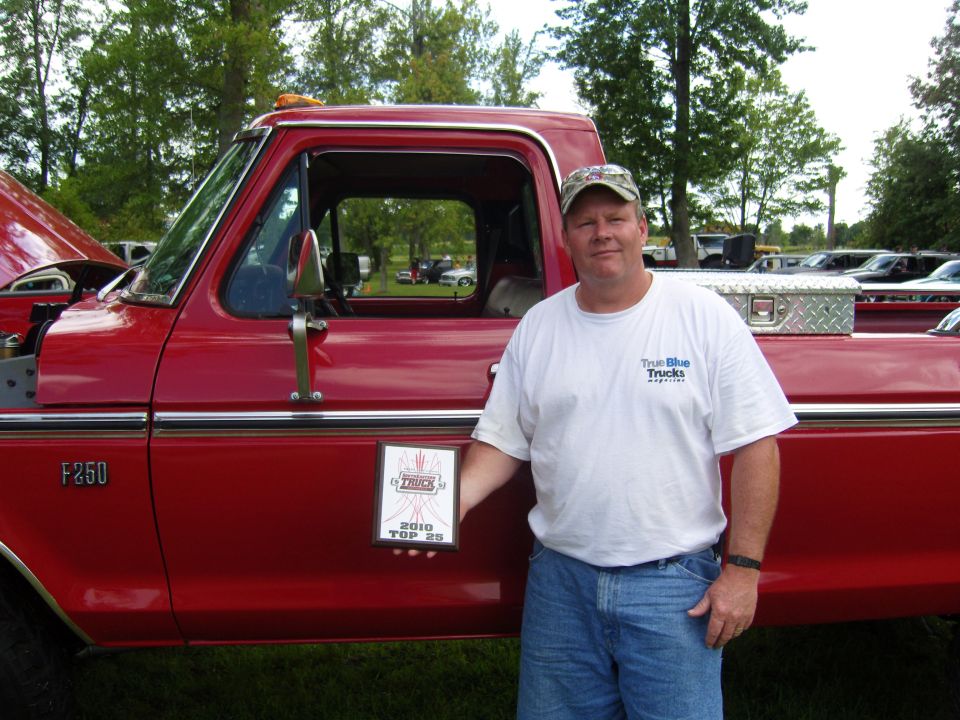 Man in white t-shirt and jeans holds a "2010 Top 25" award next to a red 1973 Ford F-250 truck. Other vehicles are parked in the background