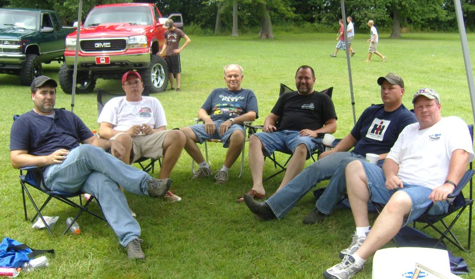 Group of men sitting in lawn chairs on grass, with two lifted trucks in the background