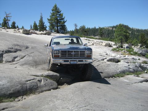 1986 Ford Bronco - This blue Ford F-Series truck, likely from the early 1980s, is shown tackling a challenging off-road course on a sunny day.