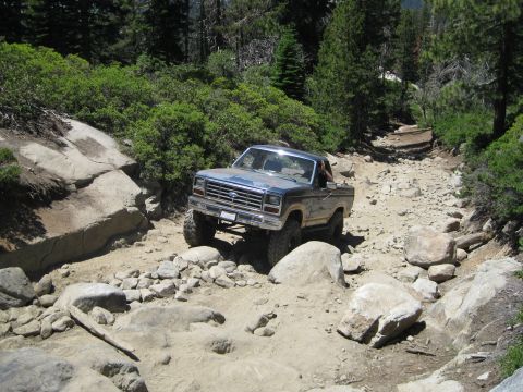 1986 Ford Bronco - A blue Ford pickup truck drives over a challenging rocky terrain on a sunny day.