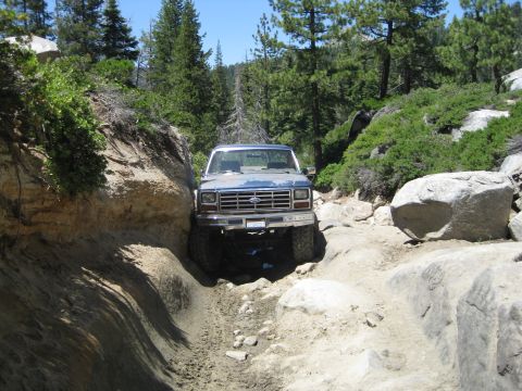 1986 Ford Bronco - Ford pickup truck is shown off-roading on a challenging, rocky trail with dense trees in the background.