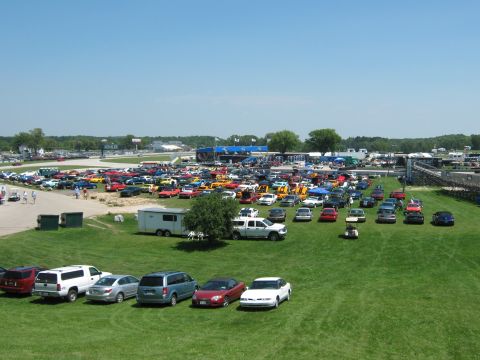 1985 Ford Mustang - A large gathering of vehicles are parked on a grassy area next to a race track on a sunny day.