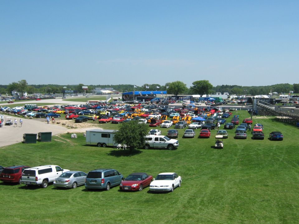 many cars parked on grass at a raceway with buildings and track in background