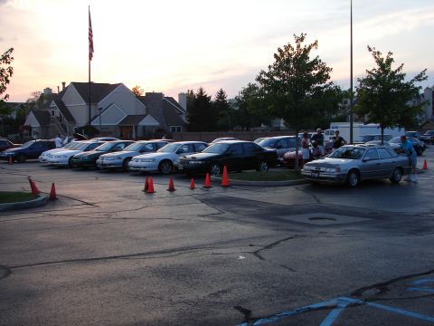 1992 Ford Taurus - Several cars are parked in a lot during an event, with an American flag flying in the background.