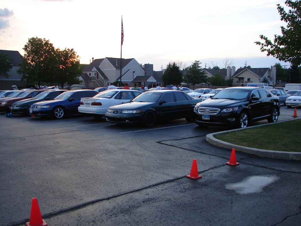 Parking lot scene with several sedans parked in rows, an American flag flying in the background