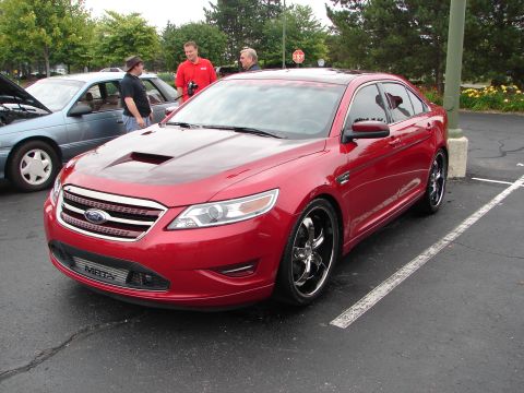 1992 Ford Taurus - A red sedan with a custom hood scoop and chrome wheels is parked in a lot, with other vehicles and people in the background.