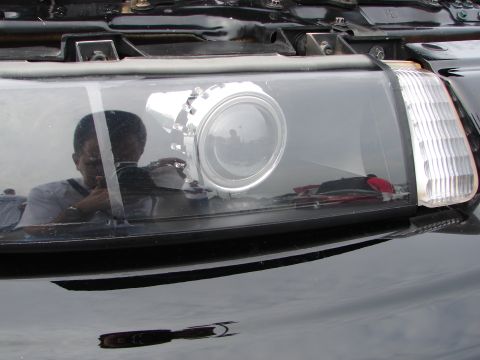 1992 Ford Taurus - A person's reflection is visible in the custom projector headlight assembly of a black vehicle.