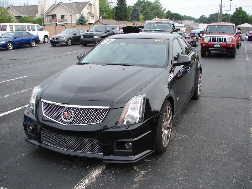 Black Cadillac CTS sedan parked in a lot, front 3/4 view. Race numbers on windshield