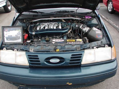1992 Ford Taurus - The engine bay of a teal car is open, revealing a V6 engine and custom modifications. A framed photo of the car sits on the fender.