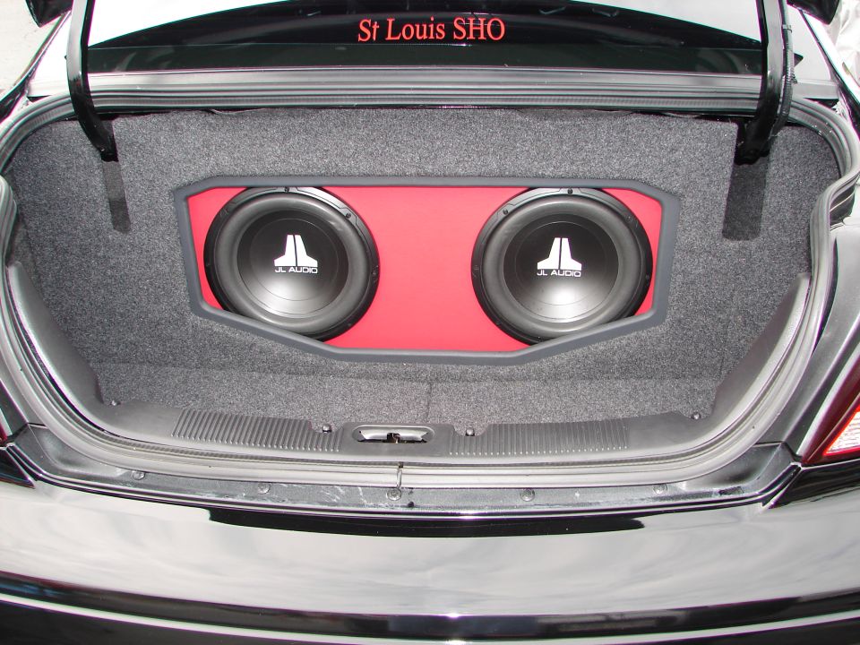 Car trunk interior with custom subwoofer box holding two JL Audio subwoofers. Red accent panel behind speakers