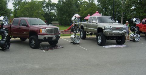 2007 Ford Mustang - Custom wheels are displayed next to lifted pickup trucks at an outdoor event.