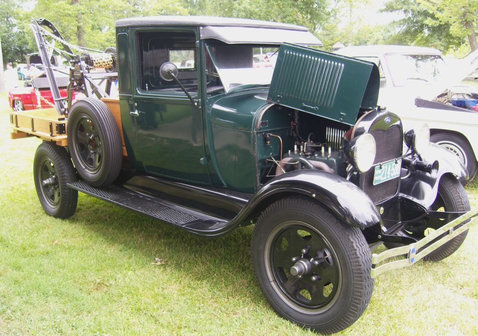 Green tow truck with a wooden bed and spare tire on the side. Hood is open showing the engine. Another white truck is partially visible behind it