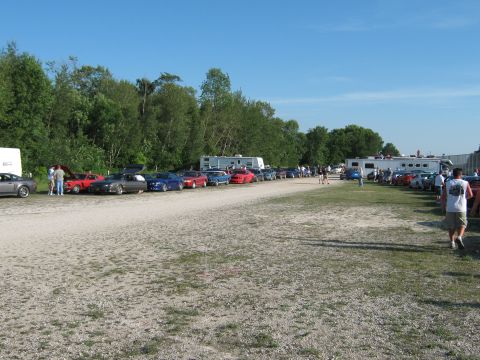 1985 Ford Mustang - A variety of cars are parked on a gravel lot under a clear blue sky, with trees and RVs visible in the background.