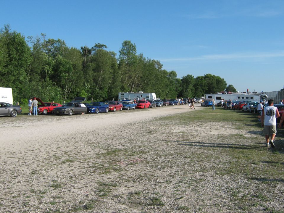 Line of cars, including a 1985 Ford Mustang, parked on a gravel lot with trees and trailers in the background