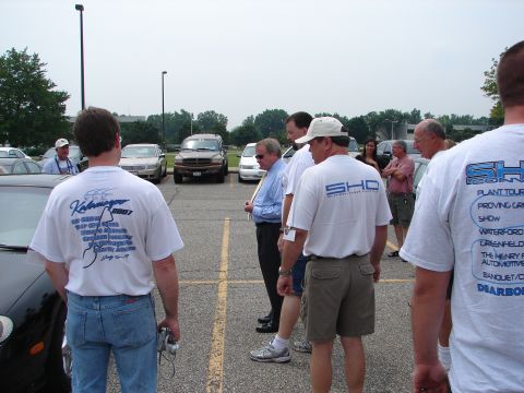 1992 Ford Taurus - A group of men wearing event t-shirts gather in a parking lot, with several cars visible in the background.