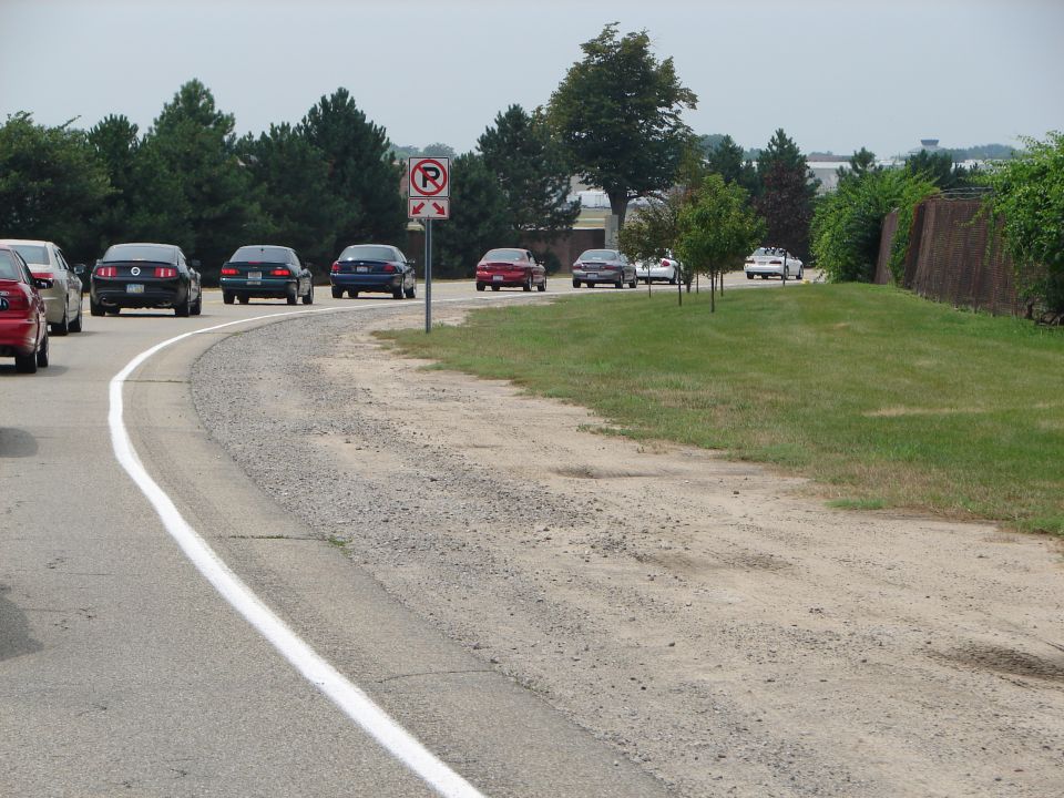 Cars driving on a road with trees and grass on the side. A no parking sign is visible
