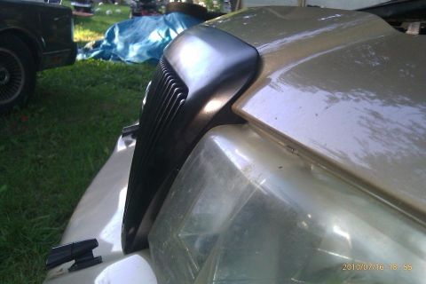2000 Mercury Grand Marquis - A close-up of the front grille and headlight of a gold 2000 Mercury Grand Marquis, with another vehicle visible in the background.