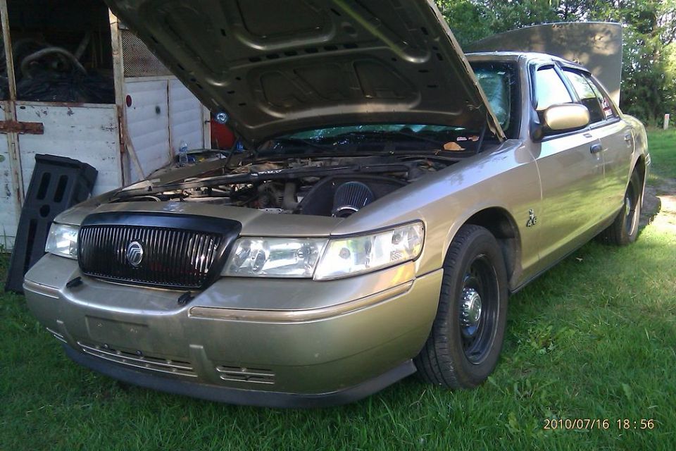Gold Mercury Grand Marquis with hood open, showing engine and aftermarket air intake. Black grille with Mercury logo