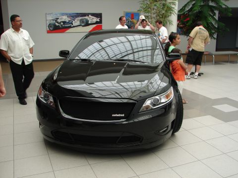 1992 Ford Taurus - A black sedan with a custom grille is displayed indoors, with people observing.