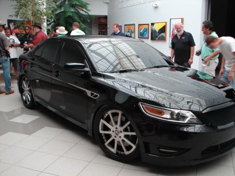 1992 Ford Taurus - A black sedan with custom wheels and a body kit is on display indoors.