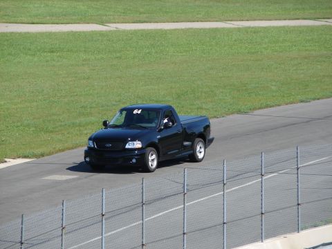 1992 Ford Taurus - A black Ford F-150 SVT Lightning pickup truck with racing number 64 is seen on a paved track with grassy areas in the background.