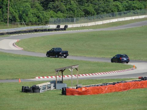 1992 Ford Taurus - A black pickup truck and a dark SUV navigate a turn on a race track with grassy infield and safety barriers.
