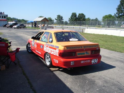 1992 Ford Taurus - A 1992 Ford Taurus, adorned with racing livery, sits on a track with a clear blue sky overhead.