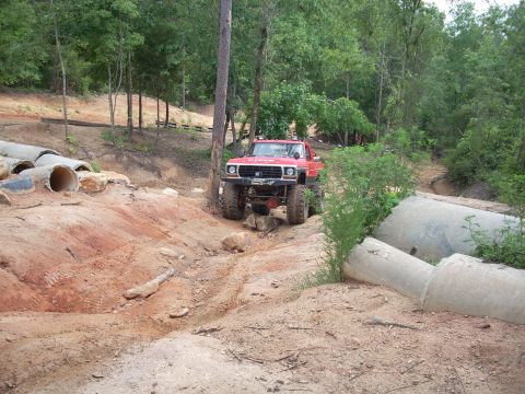 1979 Ford Bronco - A modified red Ford pickup truck drives through a challenging off-road course with large concrete pipes visible.