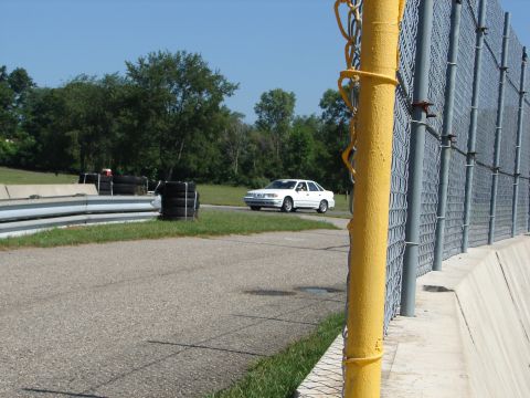 1992 Ford Taurus - A white 1992 Ford Taurus is seen on a race track with a chain-link fence and tires in the foreground.