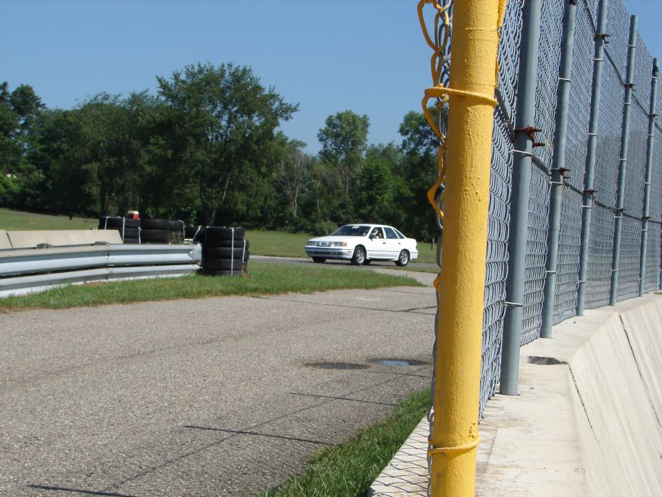 1992 Ford Taurus driving on a track, seen from the side with a chain-link fence in the foreground