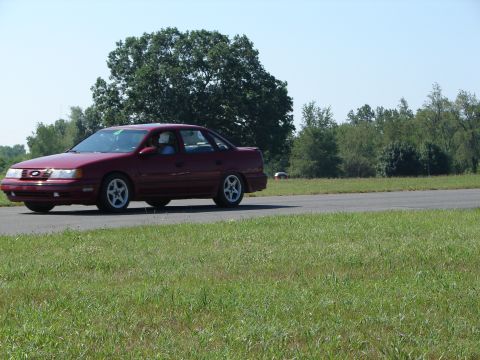 1992 Ford Taurus - A maroon sedan drives on a paved road with grassy shoulders under a clear blue sky.
