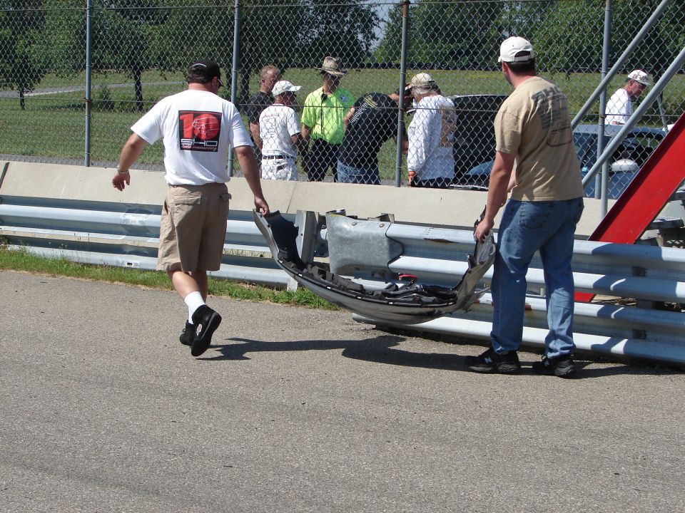 Two men carry a damaged car bumper near a guardrail at a track