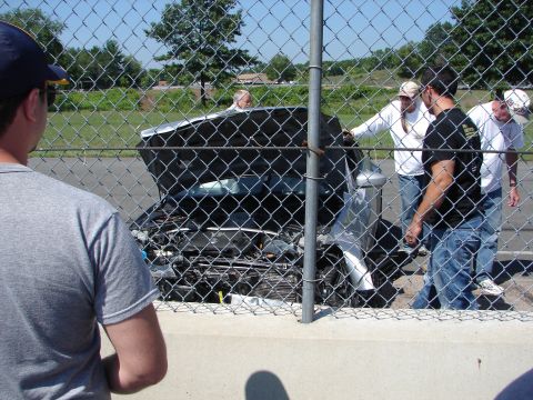 1992 Ford Taurus - People examine a damaged silver car with its hood open, viewed through a chain-link fence.