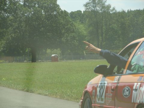 1992 Ford Taurus - A hand points from the window of a race car, with a portable toilet visible in the background.