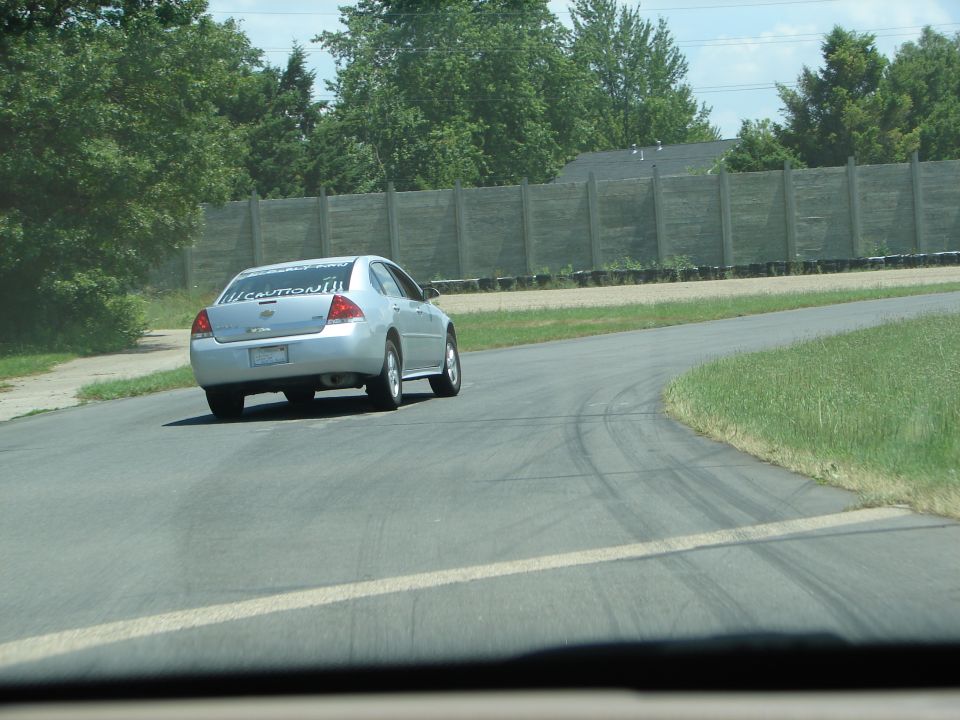 Silver sedan with 'CAUTION!!!' sticker on rear window driving on a track