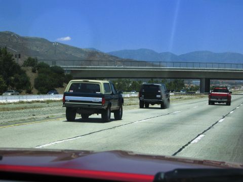1986 Ford Bronco - Three SUVs, including a 1986 Ford Bronco, travel down a highway under a bridge.