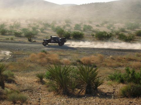 1986 Ford Bronco - A desert race truck speeds along a dusty road, surrounded by arid vegetation and hills.