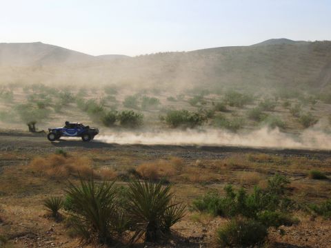 1986 Ford Bronco - An off-road buggy speeds along a dirt track, kicking up a cloud of dust in a desert landscape.