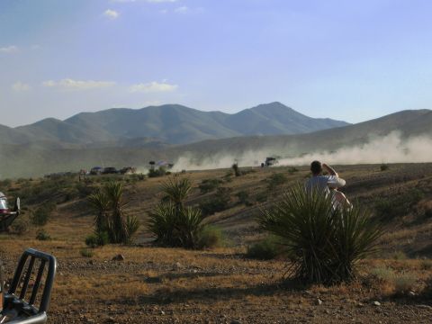 1986 Ford Bronco - A group of vehicles races across a dusty desert terrain, leaving plumes of dust in their wake under a clear sky.