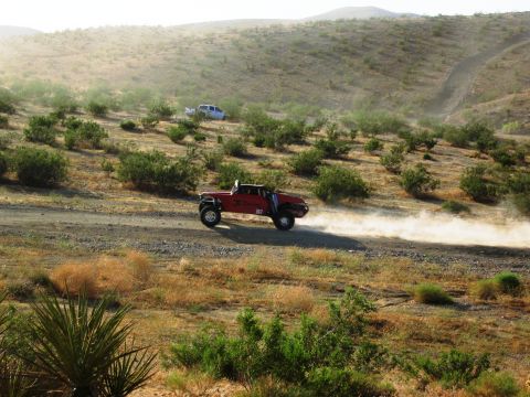 1986 Ford Bronco - A red off-road truck speeds along a dusty desert track, with another white vehicle visible in the background.