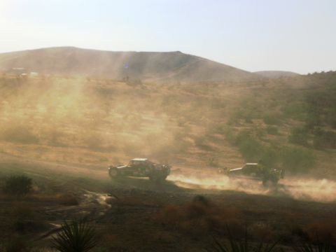 1986 Ford Bronco - Off-road trucks race on a dusty hillside under a hazy sky.