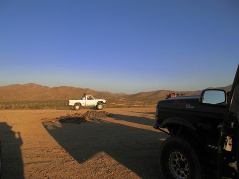 1986 Ford Bronco - Two trucks are parked in a desert area with mountains in the distance under a clear blue sky.