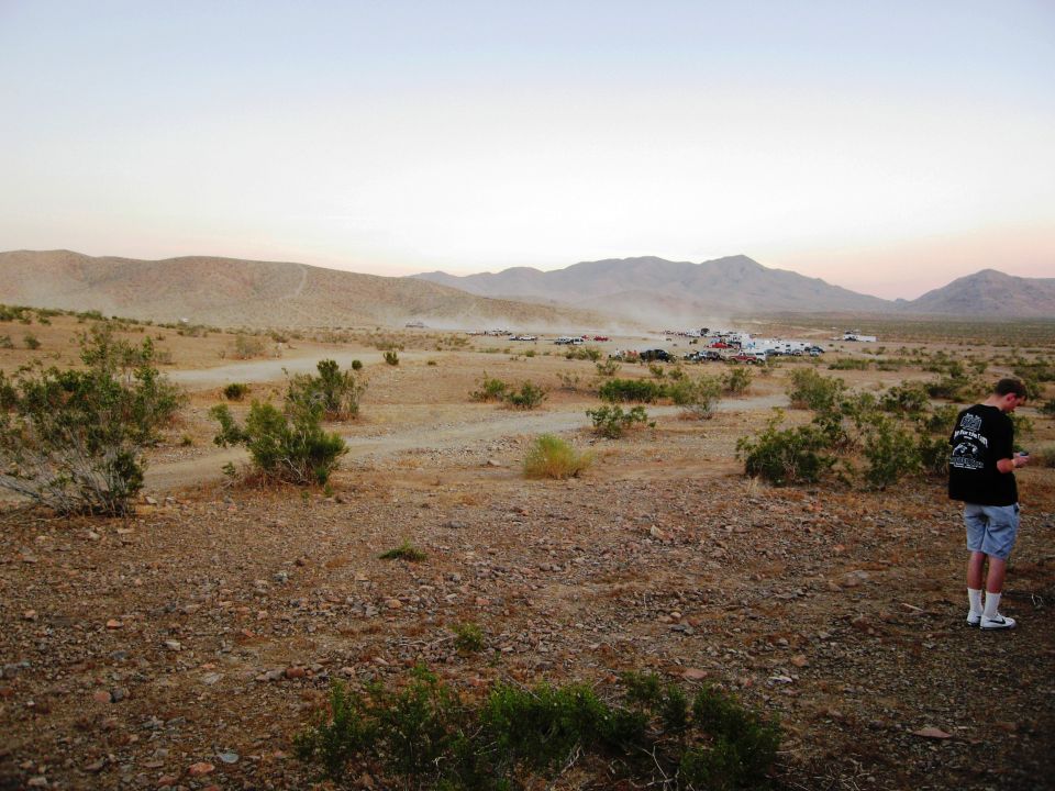 Desert scene with distant vehicles and a man in the foreground, with 'SUPERMOTORS' text at the bottom right