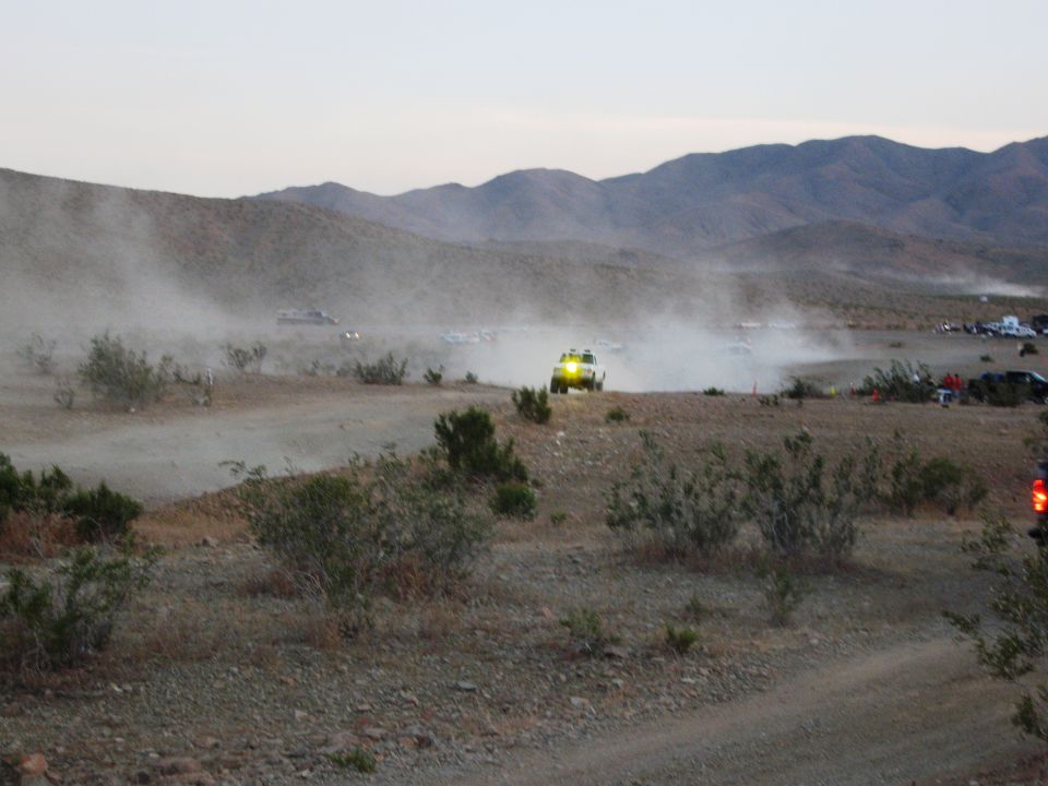 Off-road race with a yellow vehicle kicking up dust in the desert, other vehicles and spectators in background
