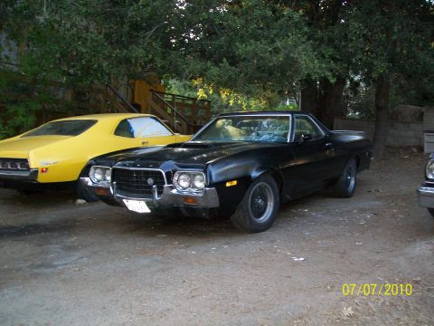 1972 Ford Ranchero - A black 1972 Ford Ranchero is parked next to a yellow Ford Torino, with trees and a wooden structure in the background.