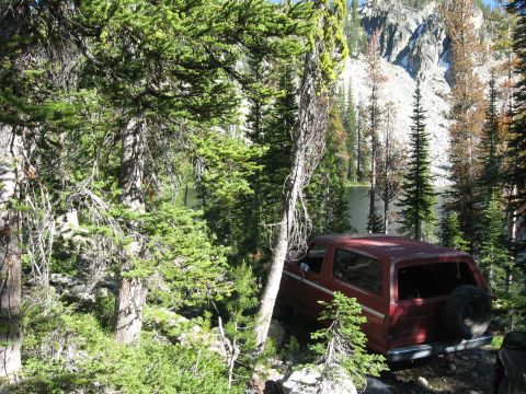 1981 Ford Bronco - A red SUV is parked off-road in a wooded area with a lake and mountains in the background.